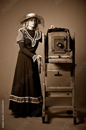 Vintage retro style photo of a young woman in a long modest dress and hat standing near a daguerreotype vintage camera on a gray background. Retro photography and vintage concept. photo