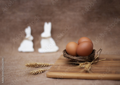 Easter decoration with eggs in the nest with spikes of wheat near bright striped board and two out of focus white rabbits on sackcloth background. Close up, copy space, horizontal. photo