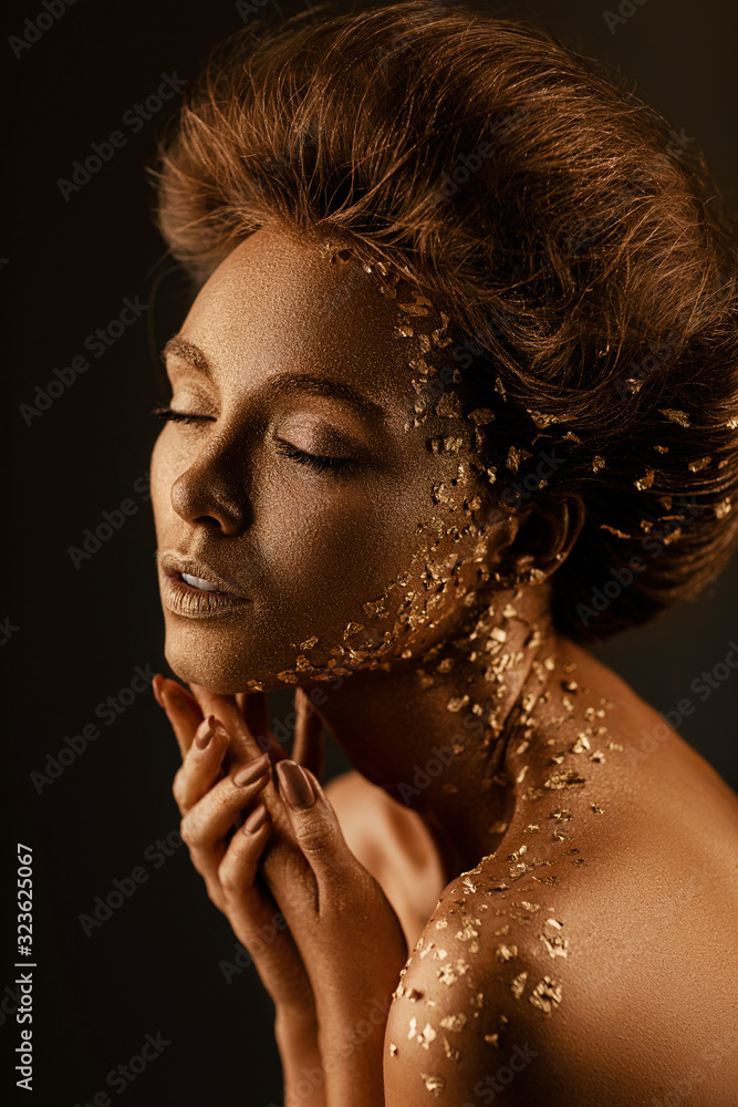 Fototapeta Fashion art portrait of model girl with holiday golden shiny professional makeup. beaty woman with golden sparkles on skin and hair on dark background. Gold glowing skin.