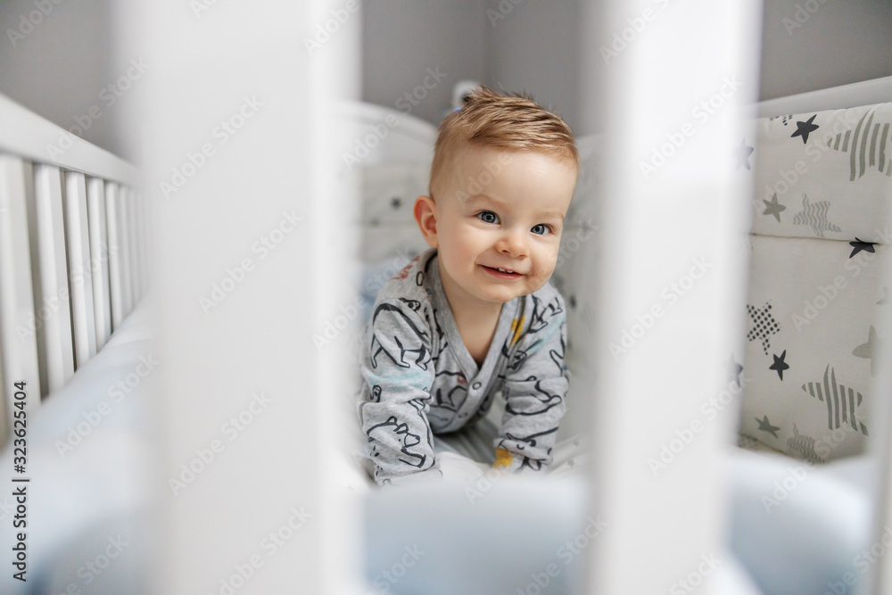 Cute blond smiling little baby boy lying on stomach in his crib and curiously looking at something interesting. Cute blond smiling little baby boy lying on stomach in his crib and curiously looking at something interesting.