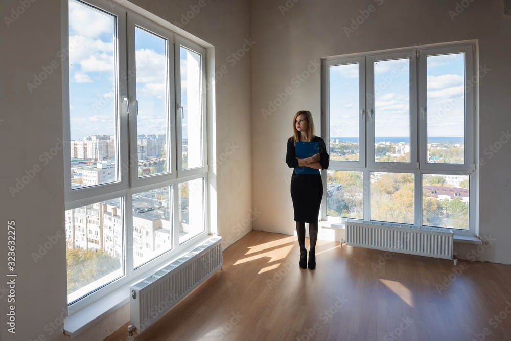 Girl realtor with a folder stands at the big windows of a spacious apartment Girl realtor with a folder stands at the big windows of a spacious apartment