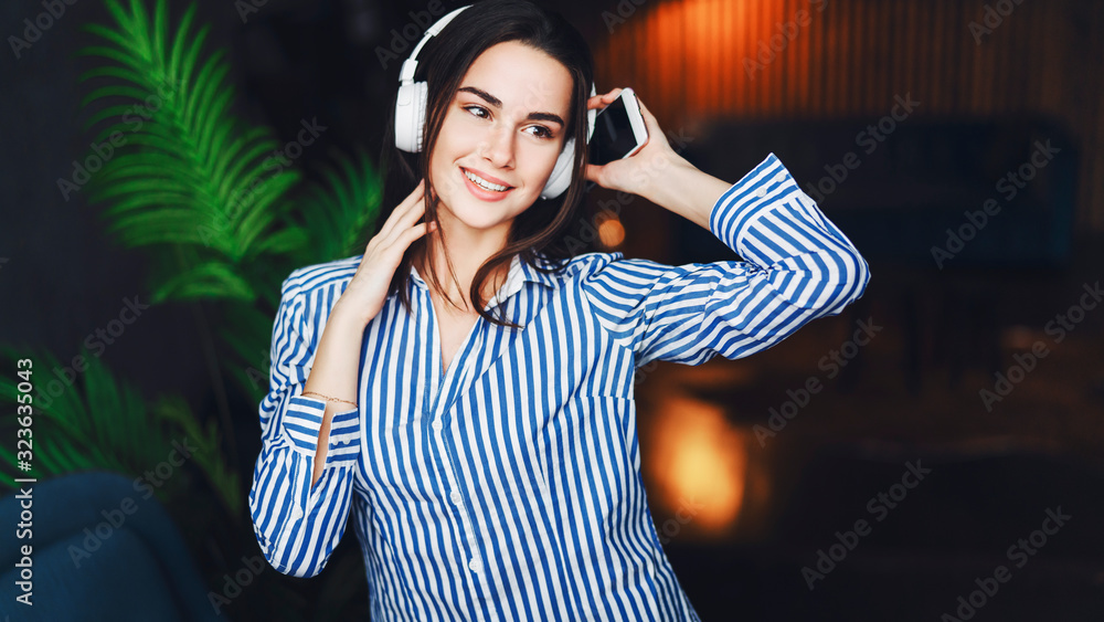 Young smiling woman standing in cafe, looking out window, holding smartphone, listening to music on headphones. Girl using gadget. Close-up of a smiling brunette in white headphones. Young smiling woman standing in cafe, looking out window, holding smartphone, listening to music on headphones. Girl using gadget. Close-up of a smiling brunette in white headphones.