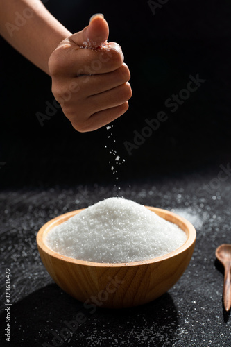 People are pouring sugar or salt to put in a wooden bowl. Black background.