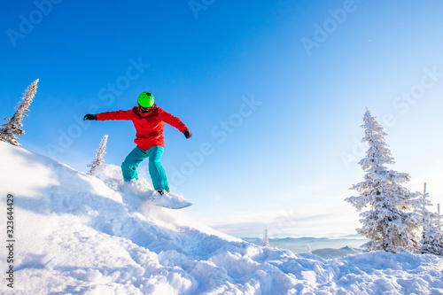Young man snowboarder jump down slope mountains forest. Winter sport sun day, blue sky, fast speed dust snow