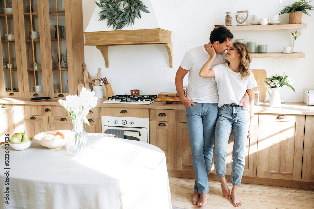 Stylishly dressed couple in love stands in the kitchen and hugging. They are wearing in jeans and white t-shirts Stylishly dressed couple in love stands in the kitchen and hugging. They are wearing in jeans and white t-shirts