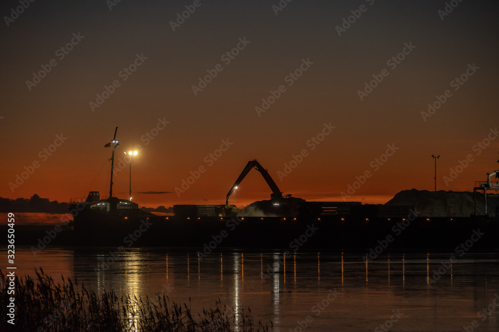 Silhouette of working crane in sea port near Parnu bay in cold winter evening against background of clear sunset sky. Port lights reflecting in water and reeds in foreground 