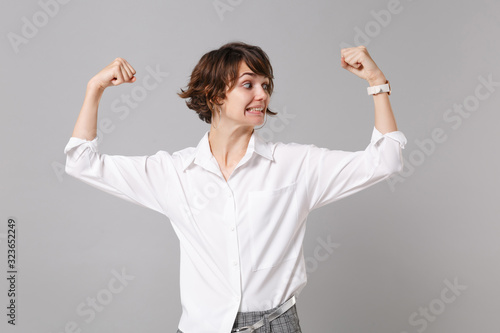 Confused amazed young business woman in white shirt posing isolated on grey wall background studio portrait. Achievement career wealth business concept. Mock up copy space. Showing biceps, muscles.