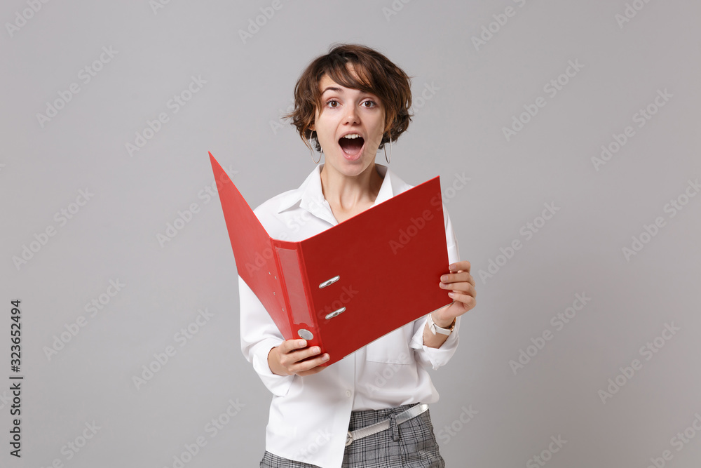 Obraz premium Excited young business woman in white shirt posing isolated on grey background studio portrait. Achievement career wealth business concept. Mock up copy space. Holding red folder for papers document.