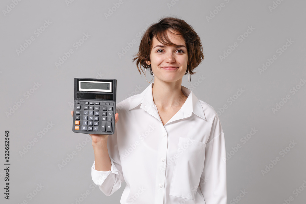 Smiling beautiful young business woman in white shirt posing isolated on grey wall background studio portrait. Achievement career wealth business concept. Mock up copy space. Hold in hand calculator. Smiling beautiful young business woman in white shirt posing isolated on grey wall background studio portrait. Achievement career wealth business concept. Mock up copy space. Hold in hand calculator.