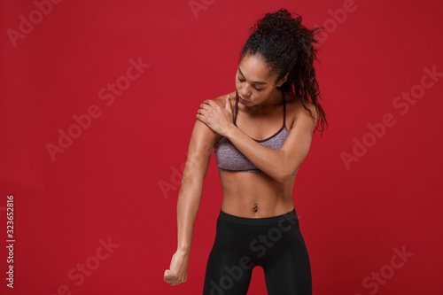 Strong young african american sports fitness woman in sportswear posing working out isolated on red wall background studio portrait. Sport exercises healthy lifestyle concept. Touching her shoulder.
