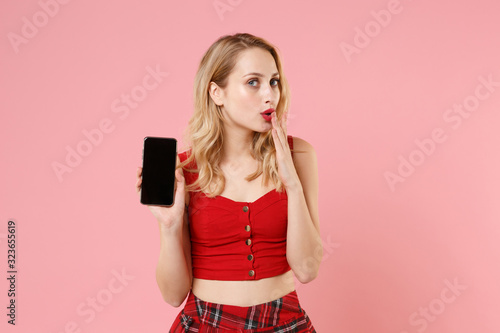 Amazed young woman in red sexy clothes posing isolated on pastel pink wall background. People lifestyle concept. Mock up copy space. Hold mobile phone with blank empty screen covering mouth with hand.