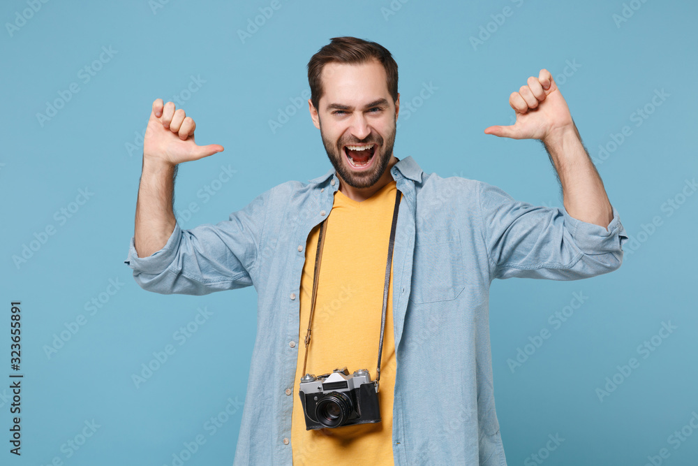 Naklejka premium Funny traveler tourist man in yellow summer clothes with photo camera isolated on blue background. Male passenger traveling abroad on weekends. Air flight journey concept. Pointing thumbs on himself.