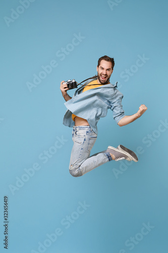 Happy traveler tourist man in yellow casual clothes with photo camera isolated on blue background. Male passenger traveling abroad on weekend. Air flight journey concept. Jumping doing winner gesture.