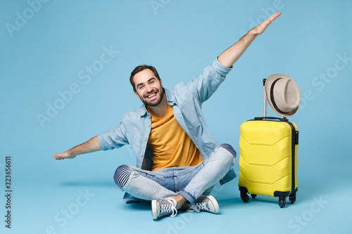 Smiling traveler tourist man in yellow clothes isolated on blue wall background. Male passenger traveling abroad on weekend. Air flight journey concept. Sit near suitcase, spreading hands like flying.
