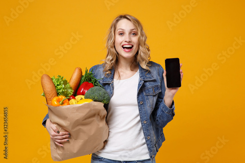 Funny girl in denim clothes isolated on orange background. Delivery service from shop or restaurant concept. Hold brown craft paper bag for takeaway with food product mobile phone with blank screen.