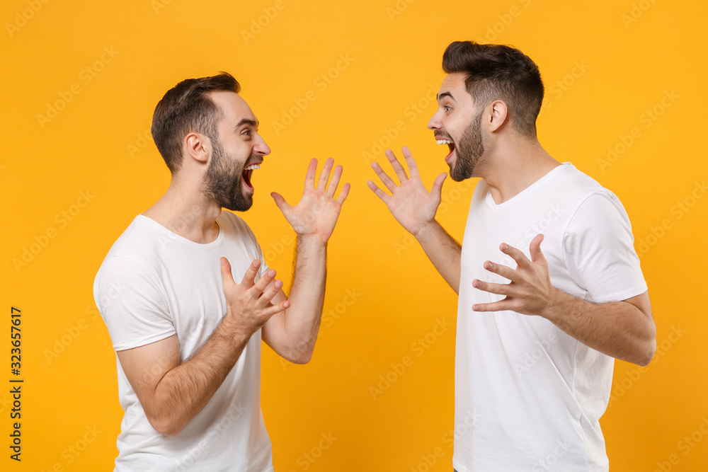 Excited young men guys friends in white blank empty t-shirts posing isolated on yellow orange background in studio. People lifestyle concept. Mock up copy space. Looking at each other spreading hands. Excited young men guys friends in white blank empty t-shirts posing isolated on yellow orange background in studio. People lifestyle concept. Mock up copy space. Looking at each other spreading hands.