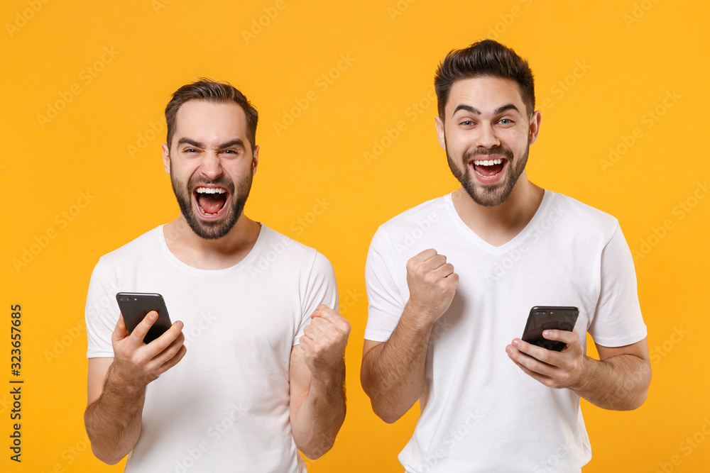 Joyful young men guys friends in white blank t-shirts isolated on yellow orange background. People lifestyle concept. Mock up copy space. Using mobile phone typing sms message doing winner gesture. Joyful young men guys friends in white blank t-shirts isolated on yellow orange background. People lifestyle concept. Mock up copy space. Using mobile phone typing sms message doing winner gesture.