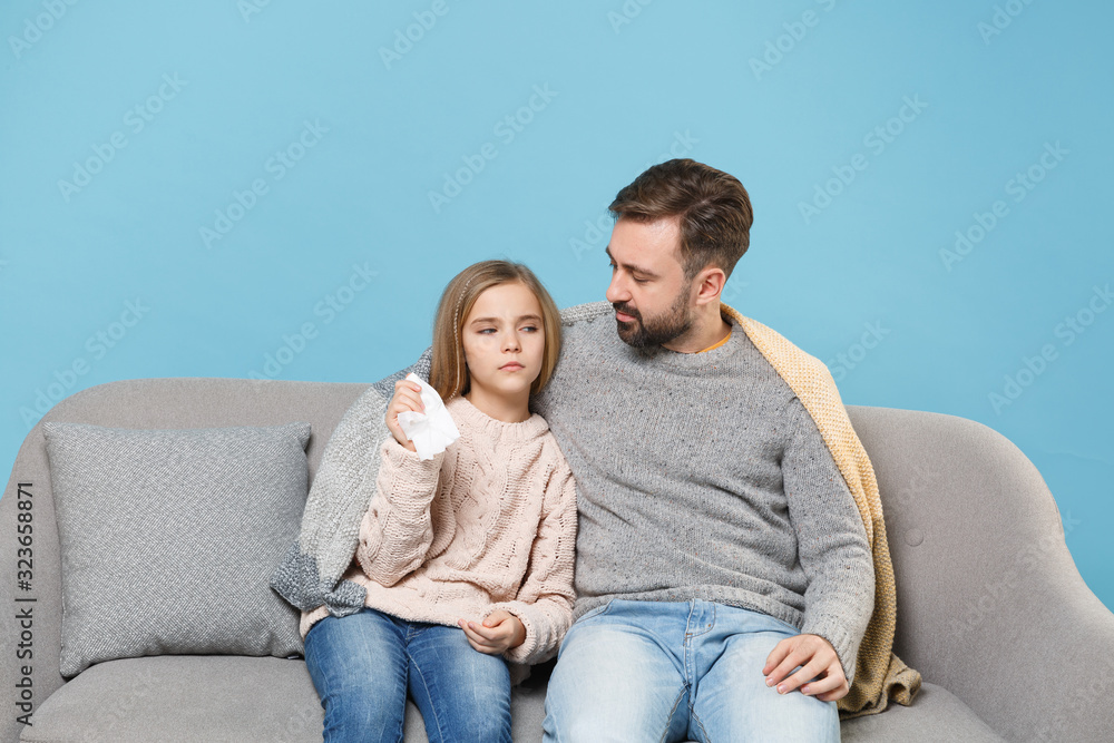Man in knitted sweater with sick child baby girl. Father little daughter isolated on blue background. Love family parenthood childhood concept. Sitting on couch, having runny nose, hold paper napkin. Man in knitted sweater with sick child baby girl. Father little daughter isolated on blue background. Love family parenthood childhood concept. Sitting on couch, having runny nose, hold paper napkin.