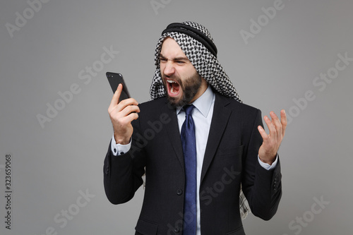 Irritated bearded arabian muslim businessman in keffiyeh kafiya ring igal agal classic black suit isolated on gray background. Achievement career wealth business concept. Talk on mobile phone scream. photo