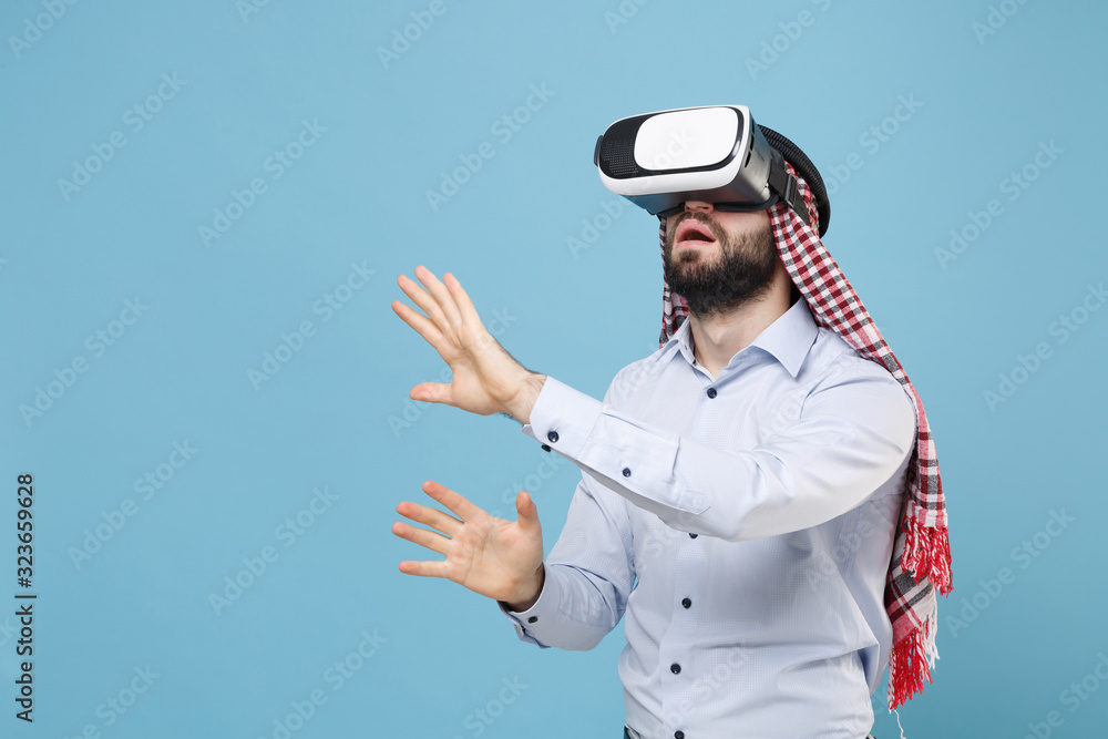 Amazed arabian muslim man in keffiyeh kafiya ring igal agal casual clothes isolated on pastel blue background. People religious lifestyle concept. Watch in headset touch something like push on button. Amazed arabian muslim man in keffiyeh kafiya ring igal agal casual clothes isolated on pastel blue background. People religious lifestyle concept. Watch in headset touch something like push on button.