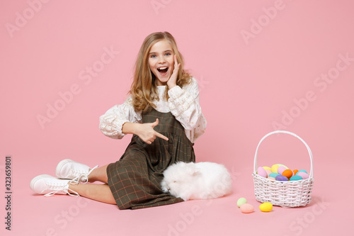Little pretty blonde kid girl 11-12 years old in light spring dress hold fluffy white bunny rabbit, wicker basket with eggs isolated on pastel pink background. Childhood lifestyle Happy Easter concept