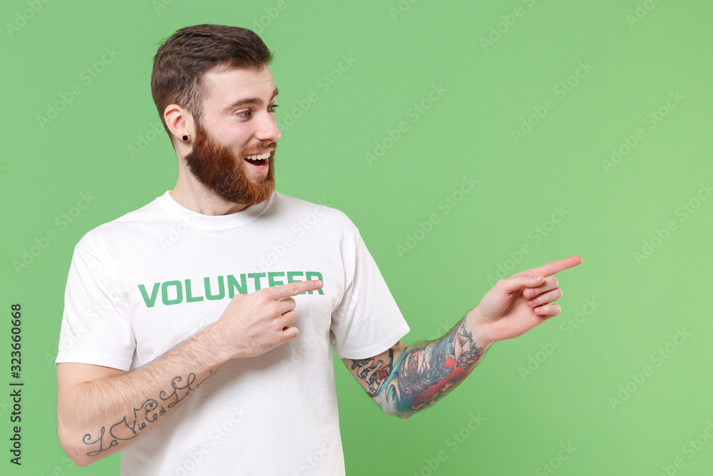 Excited young bearded man in white volunteer t-shirt isolated on pastel green background studio portrait. Voluntary free work assistance help charity grace teamwork concept. Point index fingers aside. Excited young bearded man in white volunteer t-shirt isolated on pastel green background studio portrait. Voluntary free work assistance help charity grace teamwork concept. Point index fingers aside.