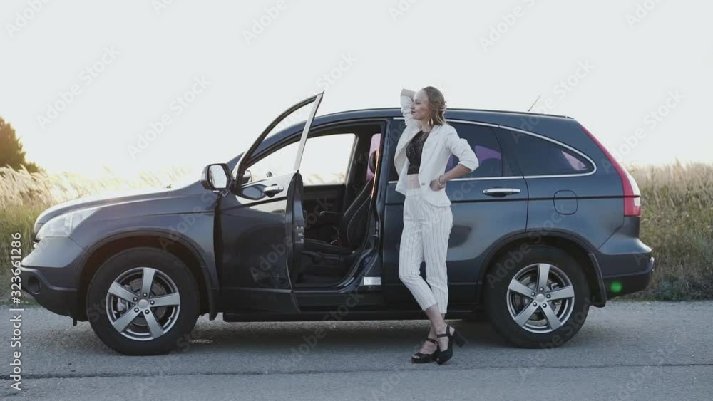 Beautiful lady in white suit and black shoes poses at car out of city Beautiful lady in white suit and black shoes poses at car out of city