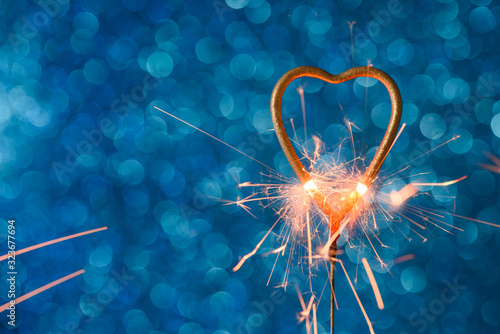 Sparkler in a shape of golden heat burning over blue bokeh background with copy-space. Wedding or Valentine's day invitation card concept.