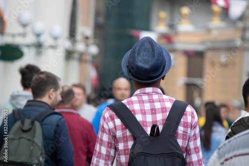 A teenager with a checkered shirt, a fedora and a backpack. The man is turned back and photographed outside. photo