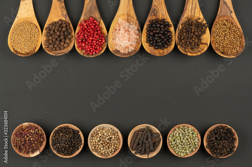 Seven cooking spoons made of olive wood in a row and six small wooden bowls in a row filled with various spices on a black background with copy space in the center