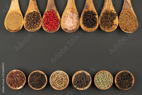 Seven cooking spoons made of olive wood in a row and six small wooden bowls in a row with various spices on a black background with copy space in the center