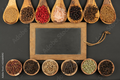 Seven cooking spoons made of olive wood in a row and six small wooden bowls in a row filled with various spices and an old black slate board with copy space on a black background