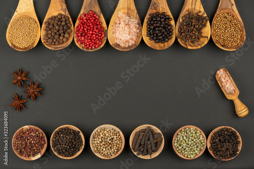Seven cooking spoons made of olive wood in a row, six small wooden bowls in a row filld with various spices on a black background with copy space in the center