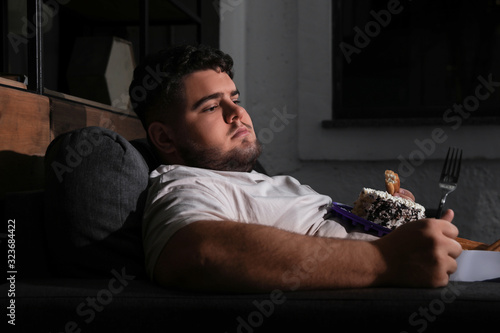 Depressed overweight man eating sweets in living room at night