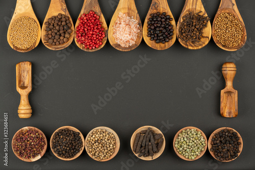 Seven cooking spoons made of olive wood in a row and six small wooden bowls in a row filled with various spices on a black background with copy space in the center