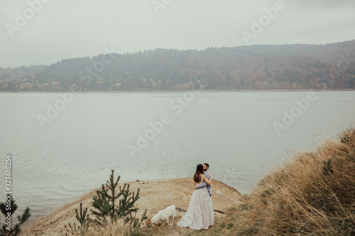 Beautiful wedding couple kissing and embracing near the shore of a mountain river.Next to the happy couple is a good white dog.Couple embracing and kissing.