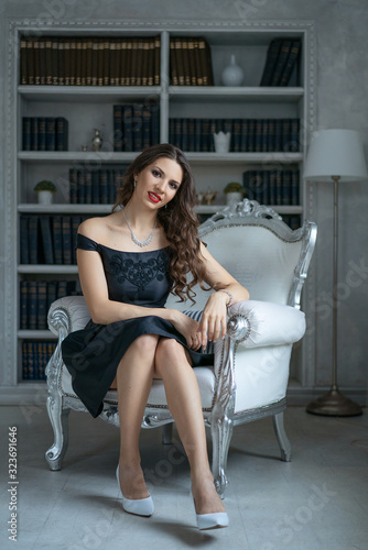 A beautiful woman with makeup and red lipstick is sitting in a black dress on a white chair against the background of shelves with books © Екатерина Переславце