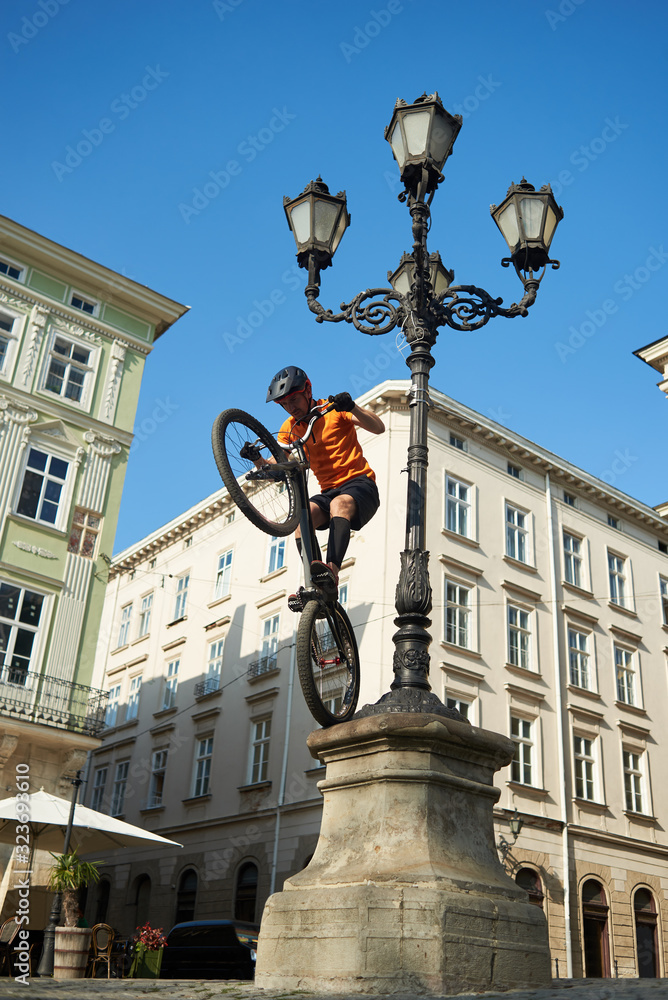 Young man is performing extremal trick with his bmx bike, keeping balance on back wheel on a pillar of ancient street lighting, low angle snapshot