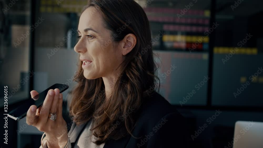 Mature businesswoman speaking into her cell phone. Business professional talking over speakerphone while standing in the office. Mature businesswoman speaking into her cell phone. Business professional talking over speakerphone while standing in the office.