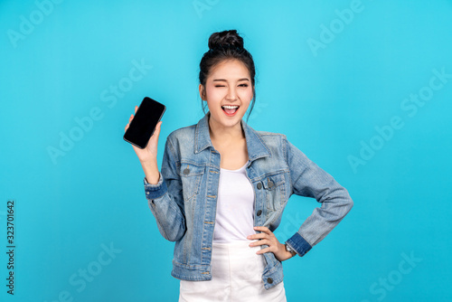 Happy asian woman feeling happiness, blinks eyes and standing hold smartphone on blue background. Cute asia girl smiling wearing casual jeans shirt and connect internet shopping online and present