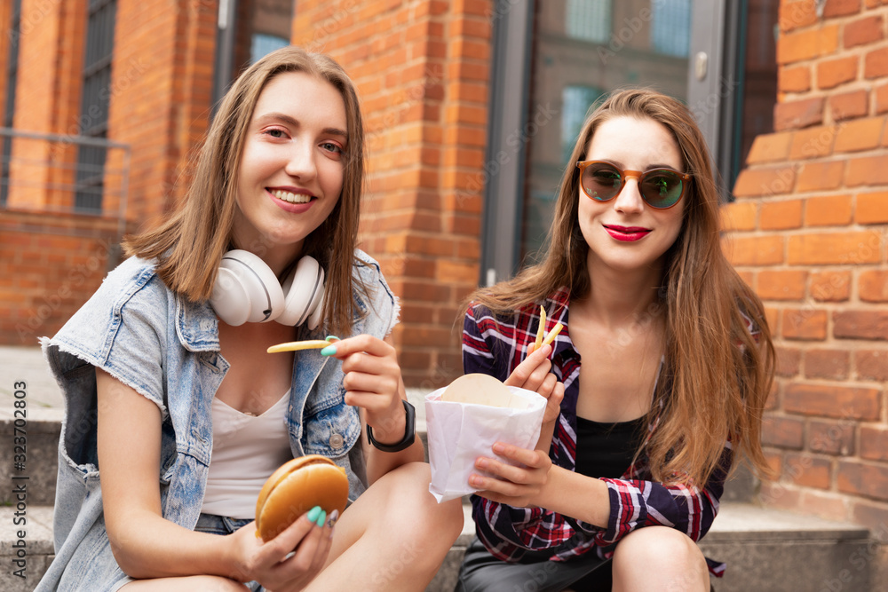 Two beautiful young girls eat fast food right on the street, they have fun, talk and feed each other delicious things.