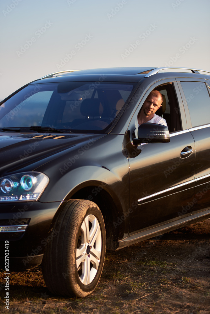 Handsome man is sitting in his black car looking out of the window smiling at the camera outside the city, glimpse of the setting sun, side view