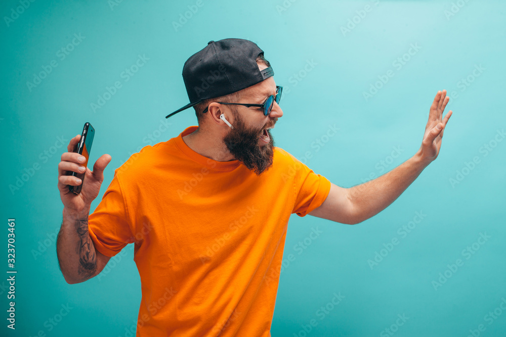 Handsome hipster guy with beard wearing orange t shirt and black cap closes eyes listens loud song in headphones, dances in rhythm of melody. Relax concept. Handsome hipster guy with beard wearing orange t shirt and black cap closes eyes listens loud song in headphones, dances in rhythm of melody. Relax concept.