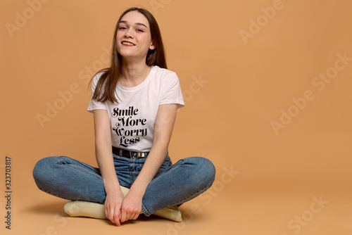 Happy female in white t-shirt sitting on the ground in lotus position and looking at the camera, orange wall with copyspace on the background