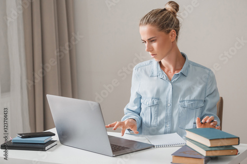 Young business woman in denim clothes freelancer working on computer keyboard typing text next to books and notebook. Office worker makes an abstract of new material. Working at a computer with papers