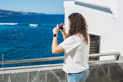 Woman taking photos with her smartphone to a summer landscape with sea and white houses lit by the sun.