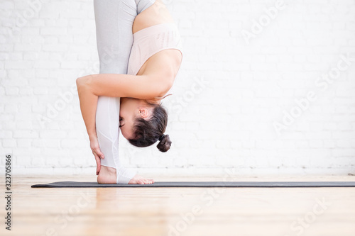 Young slim woman yoga instructor does head to knees exercise uttanasana standing forward bend pose in spacious room with large panoramic window. Concept of yoga and stretching. Advertising space photo