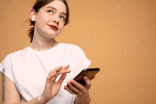 Young business lady in white t-shirt holding mobile phone in hands, wearing wireless headphones, looking up, isolaetd over orange background
