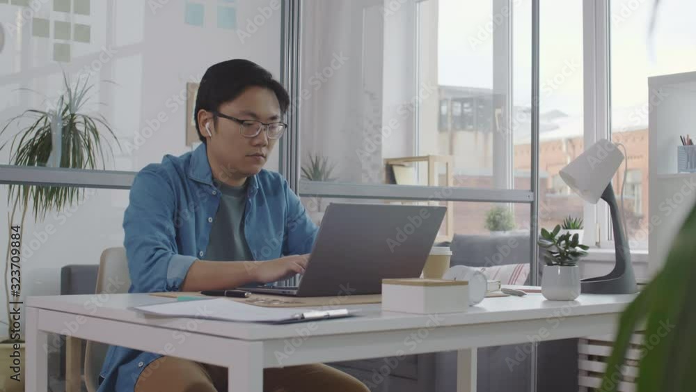 Young Asian man entering office, sitting at desk, working on laptop, drinking coffee from disposal cup, turning on table lamp and taking notes in notepad Young Asian man entering office, sitting at desk, working on laptop, drinking coffee from disposal cup, turning on table lamp and taking notes in notepad