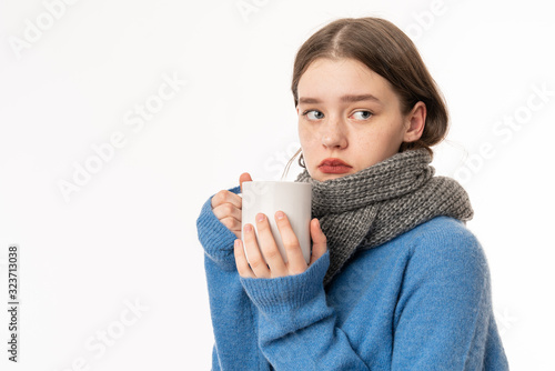 Flu treatment. Portrait of sick brunette teenage girl wearing casual clothes and scarf  holding cup of tea  studio shot isolated on white background